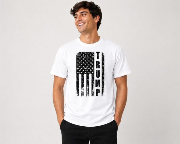 A man wearing a white short-sleeve t-shirt with a distressed black American flag graphic and the word 'TRUMP' printed vertically on the front, paired with black pants, against a light gray studio background.